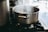 A stainless steel pot boiling with steam rising on a gas stove in a kitchen setting. The pot is prominently placed, while the background features blurred kitchen elements.