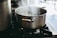 A stainless steel pot boiling with steam rising on a gas stove in a kitchen setting. The pot is prominently placed, while the background features blurred kitchen elements.
