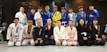 A group of people dressed in martial arts uniforms, likely participating in a Brazilian Jiu-Jitsu class, posed together for a group photo in a dojo or gym with exercise equipment in the background. The mix of uniforms includes various colors such as white, blue, black, pink, and yellow.