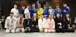 A group of people dressed in martial arts uniforms, likely participating in a Brazilian Jiu-Jitsu class, posed together for a group photo in a dojo or gym with exercise equipment in the background. The mix of uniforms includes various colors such as white, blue, black, pink, and yellow.