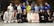 A group of people dressed in martial arts uniforms, likely participating in a Brazilian Jiu-Jitsu class, posed together for a group photo in a dojo or gym with exercise equipment in the background. The mix of uniforms includes various colors such as white, blue, black, pink, and yellow.