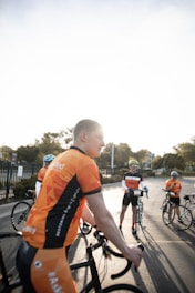 Several cyclists are gathered on a road wearing matching orange and black cycling jerseys. Each person appears to be preparing for a ride, holding their bicycles. The light from the sunrise or sunset creates a bright, warm atmosphere.