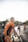 Several cyclists are gathered on a road wearing matching orange and black cycling jerseys. Each person appears to be preparing for a ride, holding their bicycles. The light from the sunrise or sunset creates a bright, warm atmosphere.