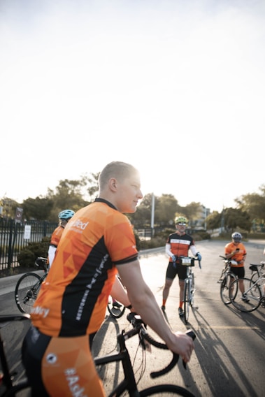 Several cyclists are gathered on a road wearing matching orange and black cycling jerseys. Each person appears to be preparing for a ride, holding their bicycles. The light from the sunrise or sunset creates a bright, warm atmosphere.