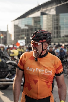 A person dressed in an orange cycling jersey and helmet is smiling in a lively urban setting. Other cyclists and motorcycles are in the background near a modern building.