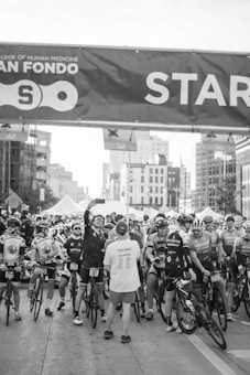 A group of cyclists gather at a starting line for an event, with many wearing cycling gear and helmets. One person stands with their back to the camera wearing a 'volunteer' shirt, while another cyclist takes a selfie. Behind them are several tall buildings and tents, indicating an urban setting for the event.