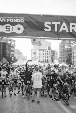 A group of cyclists gather at a starting line for an event, with many wearing cycling gear and helmets. One person stands with their back to the camera wearing a 'volunteer' shirt, while another cyclist takes a selfie. Behind them are several tall buildings and tents, indicating an urban setting for the event.