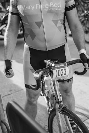 A person wearing a cycling jersey and bib shorts is holding a bicycle. The cycling jersey displays the words 'Mary Free Bed Rehabilitation'. The bicycle has a mounted number 1033. The setting appears to be an event, possibly a race or a group ride.