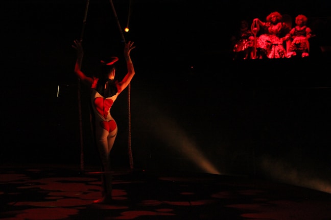 A trapeze artist is suspended in the air in a dimly lit space, illuminated by a spotlight. The artist is wearing a form-fitting costume with a bright pattern. In the background, a group of figures sits in darkness, with a red light casting a glow on them, creating a dramatic and mysterious atmosphere.