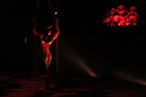 A trapeze artist is suspended in the air in a dimly lit space, illuminated by a spotlight. The artist is wearing a form-fitting costume with a bright pattern. In the background, a group of figures sits in darkness, with a red light casting a glow on them, creating a dramatic and mysterious atmosphere.
