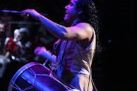 A musician is energetically playing a drum with drumsticks in a dimly lit environment. The focus is on the musician's expressive pose, with water droplets visible on his skin, suggesting intensity and physical effort. The background is blurred, indicating a crowd or audience, enhancing the dynamic atmosphere.