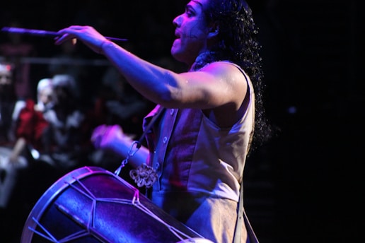 A musician is energetically playing a drum with drumsticks in a dimly lit environment. The focus is on the musician's expressive pose, with water droplets visible on his skin, suggesting intensity and physical effort. The background is blurred, indicating a crowd or audience, enhancing the dynamic atmosphere.