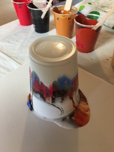 An upside-down plastic cup with multicolored paint drips on a canvas, surrounded by other cups filled with different colors of paint. The paint has spilled out from the cup, creating an intriguing mix of colors. The scene suggests a paint-pouring art activity.