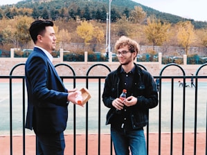 Two individuals engage in a conversation near a black fence with a sports field in the background. One wears a blue suit and holds a drink, while the other is dressed in casual black attire, holding a soda bottle. Trees with autumn foliage and a hill are visible in the distance.