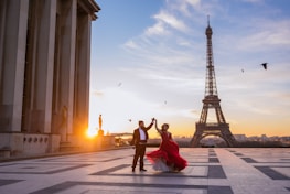 A couple dance in front of the Eiffel Tower at sunrise. The woman wears a flowing red dress, and the man is in a suit. The setting sun casts a warm glow over the scene, while birds fly in the sky.