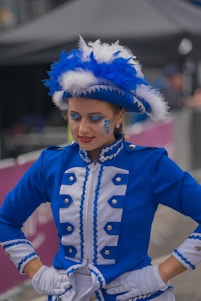 A person dressed in a vibrant blue and white costume with elaborate trimmings and matching gloves. The outfit is adorned with silver buttons and features a matching hat with prominent white and blue feathers. The person has glittery blue makeup around the eyes and a calm expression.