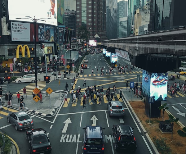 people walking on pathway near buildings and different vehicles on road under white and gray sky during daytime