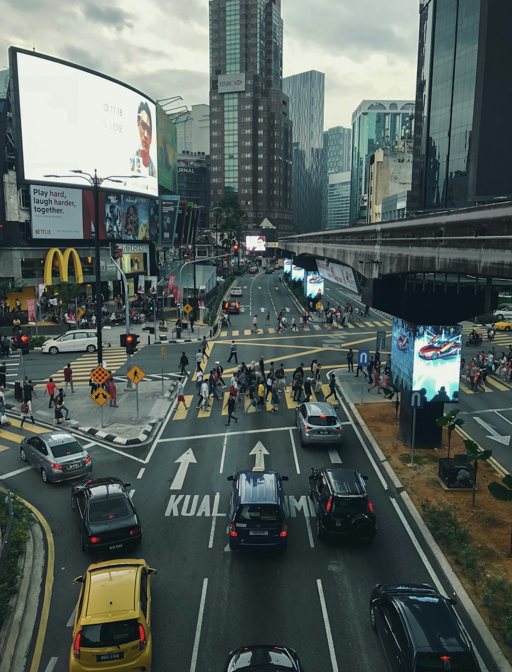 people walking on pathway near buildings and different vehicles on road under white and gray sky during daytime