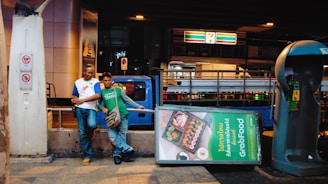 Two people stand casually next to a tilted GrabFood advertisement on a city street. Behind them, a blue truck is partially visible. A 7-Eleven sign is lit up in the background above a pedestrian walkway. A richly colored phone booth stands nearby, and the atmosphere suggests an urban setting during the evening hours.