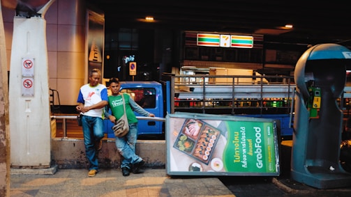 Two people stand casually next to a tilted GrabFood advertisement on a city street. Behind them, a blue truck is partially visible. A 7-Eleven sign is lit up in the background above a pedestrian walkway. A richly colored phone booth stands nearby, and the atmosphere suggests an urban setting during the evening hours.