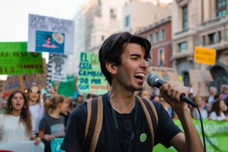 A young person passionately speaks into a microphone while participating in a public demonstration. The crowd behind them holds various signs and banners with messages, suggesting the event is a protest or march. The backdrop includes urban architecture.