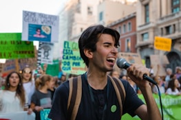 A young person passionately speaks into a microphone while participating in a public demonstration. The crowd behind them holds various signs and banners with messages, suggesting the event is a protest or march. The backdrop includes urban architecture.