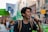 A young person passionately speaks into a microphone while participating in a public demonstration. The crowd behind them holds various signs and banners with messages, suggesting the event is a protest or march. The backdrop includes urban architecture.