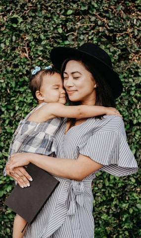 A woman wearing a striped dress and black hat holds a small child with a blue hair clip. They are embracing lovingly in front of a lush green leafy background.