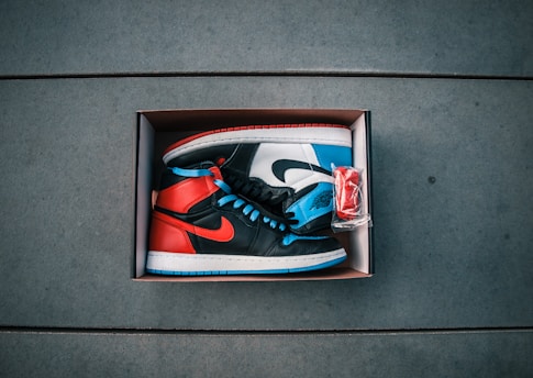 A pair of high-top sneakers is displayed inside a shoe box. The shoes have a vibrant color scheme featuring red, blue, and black panels, with matching blue laces and distinctive logos. Next to the sneakers, a small plastic bag containing an extra pair of red laces is visible. The background consists of a smooth, tiled surface.
