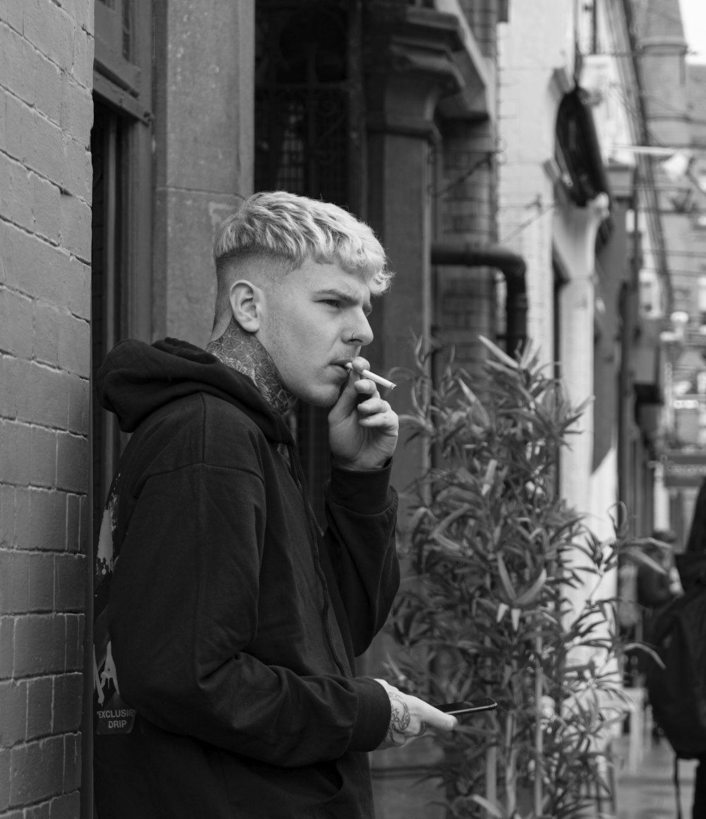 벽돌 벽에 기대어 담배를 피우는 남자의 회색조 사진