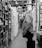 A man stands in a book aisle, intently browsing through books on a shelf in a narrow, indoor market setting. The environment is cluttered with closed metal shutters and various stalls lining the pathway, creating a confined yet inviting atmosphere for book shopping.