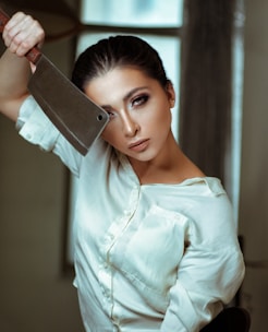 A person in a white shirt is holding a large cleaver near their face with a focused expression. The background is softly blurred, and the setting appears indoors.