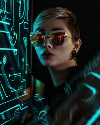 A person with short hair and reflective sunglasses stands next to a futuristic digital interface with neon blue lines. The lighting casts a dramatic effect, with shadows and bright highlights.