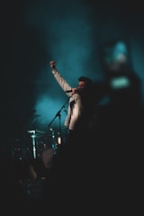 A person stands on stage, gripping a microphone and raising a fist triumphantly. The background is dark with a teal hue, suggesting a concert or performance environment. Musical instruments, including drums, are faintly visible in the background.