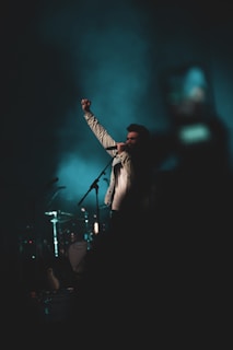 A person stands on stage, gripping a microphone and raising a fist triumphantly. The background is dark with a teal hue, suggesting a concert or performance environment. Musical instruments, including drums, are faintly visible in the background.