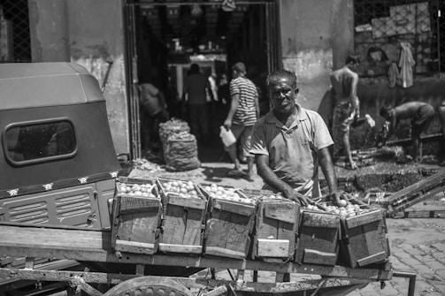 A monochromatic image of a street market scene featuring a vendor standing beside a cart filled with crates of produce. The background includes other people engaging in various activities, such as washing and transporting goods. The setting appears rustic and lively, with visible signs of daily commerce.