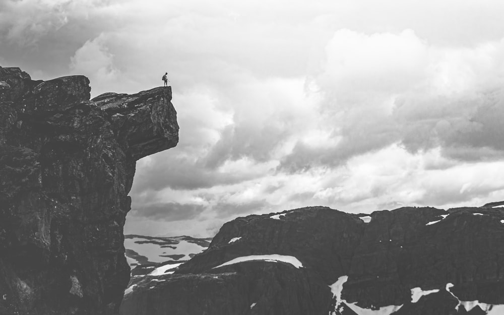 grayscale photo of person standing on rock formation under cloudy sky