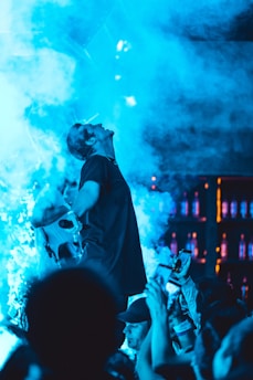 A performer is energetically engaged with a crowd in a vibrant, smoke-filled environment. Blue lighting dominates the scene, creating a dynamic and electric atmosphere. People in the audience hold up their phones, capturing the moment, as the performer gestures dramatically on stage.