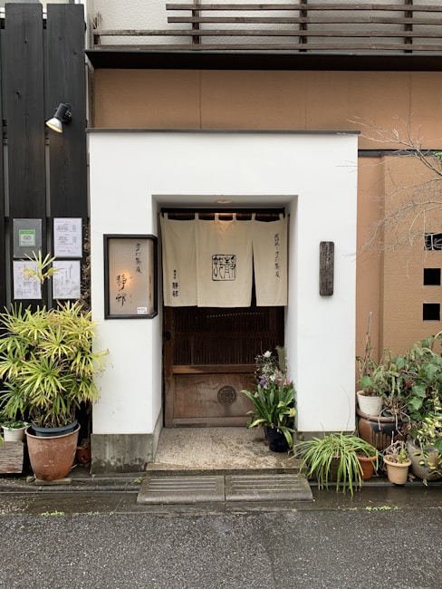 A small entrance to a traditional Japanese establishment with a white fabric banner hanging above a wooden door. The entryway is framed by various potted plants, including ferns and flowers. Wooden panels and signage with Japanese characters are visible, contributing to the quaint and authentic atmosphere.