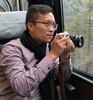 A person holding a camera is sitting next to a window on a train. The individual is intently focusing on capturing a photograph, wearing glasses, a checkered shirt, and a black scarf. Outside the window, there is a blurred view of a forested area.