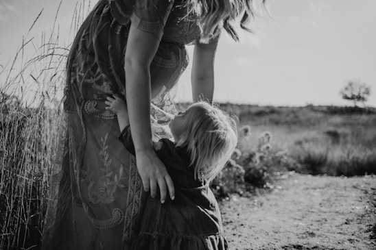 A black and white photograph capturing a young child reaching up to embrace an adult in a scenic outdoor setting. Tall grass surrounds the pair, and the adult is dressed in a textured, patterned dress. The landscape suggests a natural, open field under a partially cloudy sky.