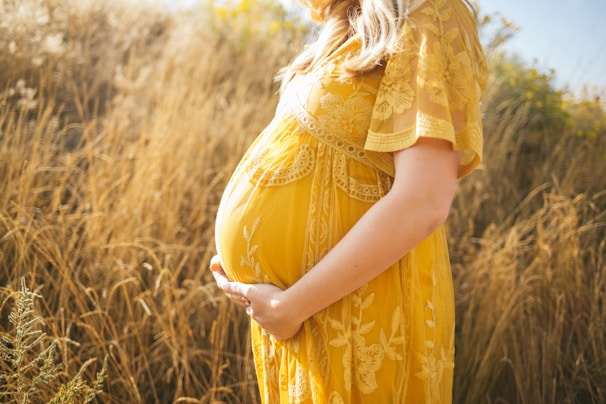 A pregnant woman is standing in a field of tall, dry grasses. She is wearing a yellow, floral-patterned dress and is gently cradling her belly with both hands. The sunlight creates a warm, serene atmosphere, highlighting the intricate details of the dress and the natural setting.