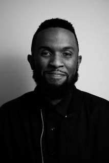 A black and white portrait of a person with a beard and short hair, smiling slightly while wearing a dark jacket and shirt. The background is plain, creating a minimalist and elegant feel.