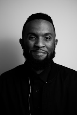 A black and white portrait of a person with a beard and short hair, smiling slightly while wearing a dark jacket and shirt. The background is plain, creating a minimalist and elegant feel.