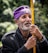 An older man with a gray beard is wearing a purple headpiece with a cross. He holds a yellow staff close to his chest. His expression appears thoughtful or reflective, and he is dressed in a dark coat. There are blurred trees in the background, suggesting an outdoor setting.