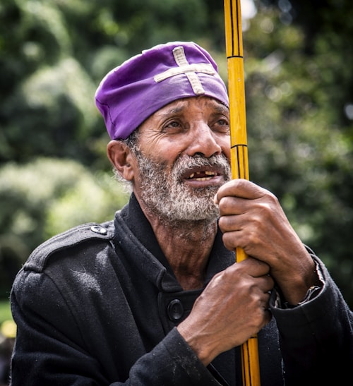An older man with a gray beard is wearing a purple headpiece with a cross. He holds a yellow staff close to his chest. His expression appears thoughtful or reflective, and he is dressed in a dark coat. There are blurred trees in the background, suggesting an outdoor setting.