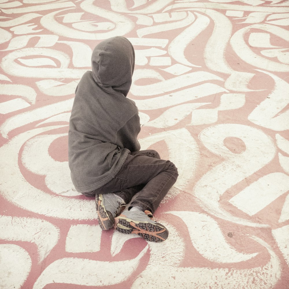 person wearing gray hooded jacket and gray denim jeans sitting on white and pink concrete pavement
