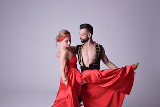 Two people are engaged in a dance, wearing elegant and vibrant costumes. The woman is dressed in a long, flowing red dress, with a matching headband holding her hair back. The man is wearing a black outfit with gold accents, featuring an open front. They are in a dynamic pose, exuding grace and coordination.