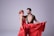 Two people are engaged in a dance, wearing elegant and vibrant costumes. The woman is dressed in a long, flowing red dress, with a matching headband holding her hair back. The man is wearing a black outfit with gold accents, featuring an open front. They are in a dynamic pose, exuding grace and coordination.