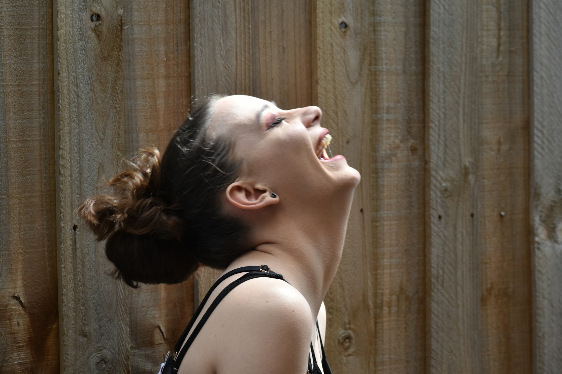 A person with their head tilted back, laughing joyfully in front of a wooden fence. The individual has dark hair pulled into a bun and wears a sleeveless top.